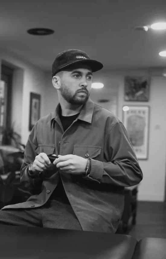 A black and white photo of Blaze, a tattoo artist, wearing a cap and jacket, standing inside a tattoo studio and looking thoughtfully to the side.