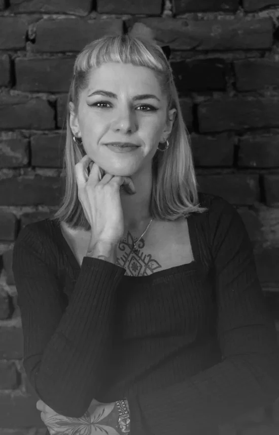 A black and white photo of Irina, a tattoo artist, posing with a hand resting on her chin, smiling confidently in front of a brick wall. Her chest tattoo is partially visible.